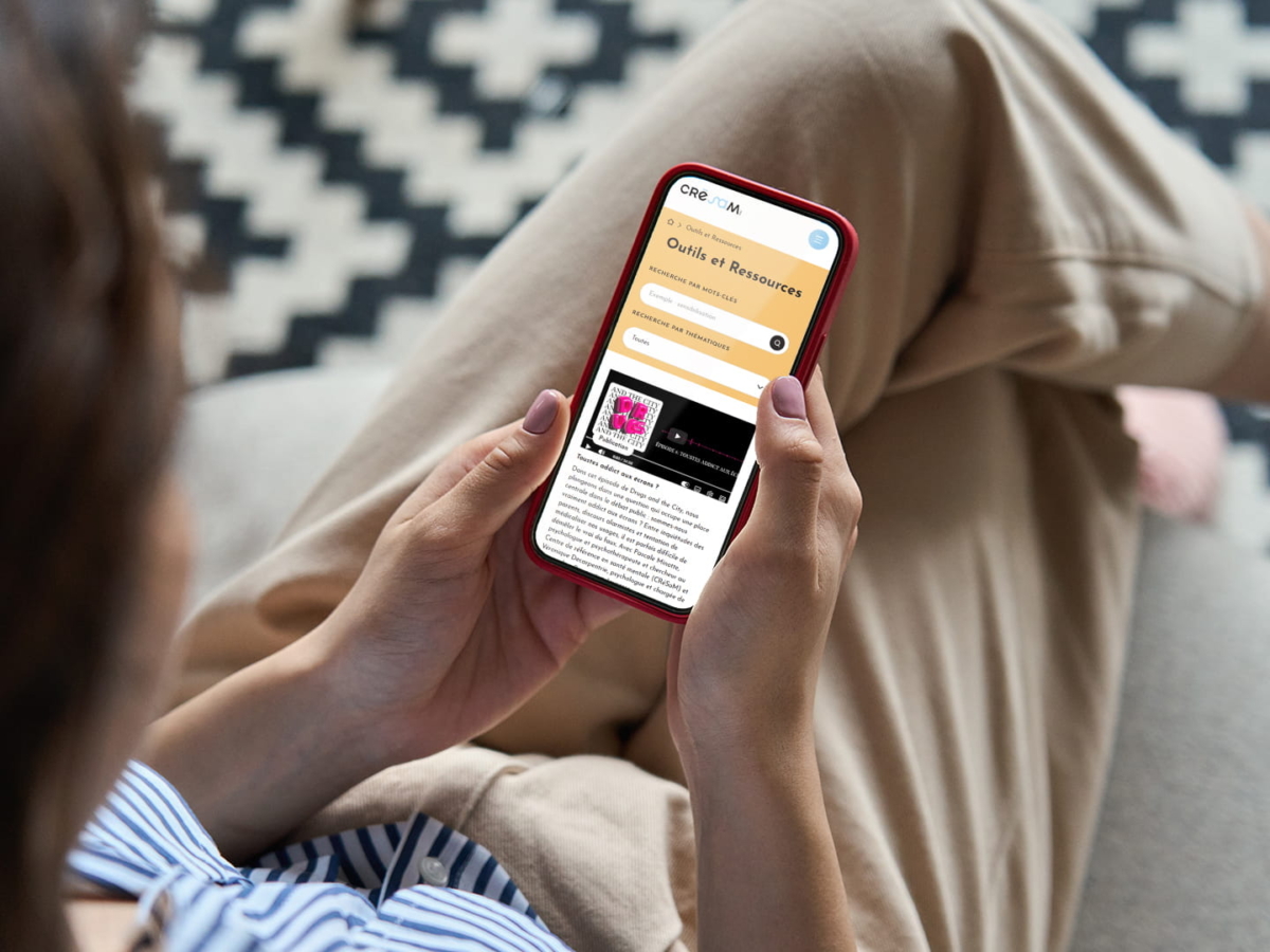 Mains d'une personne tenant un smartphone qui affiche le site mobile du CRESAM